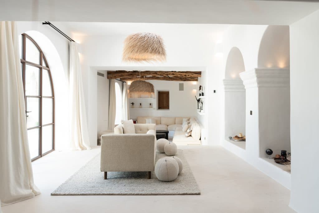 Bright, minimalist living room with white walls, arched windows, beige sofas, pouffes, textured rug, rustic wooden ceiling beam, and woven pendant lights, creating an airy, modern, and cosy atmosphere. | Ibiza