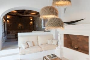 A cosy, white-walled living room with a built-in cream sofa, soft beige cushions, a modern white fireplace, rustic wood-accented ceiling, and three hanging lights with straw shades. A coffee table holds books in the foreground. | Ibiza