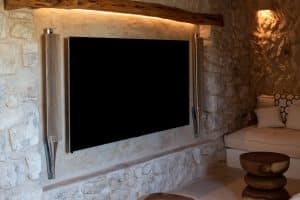 A modern flat-screen TV is mounted on a stone wall with two vertical speakers beside it. There is a built-in cushioned seating area with patterned cushions and a round wooden coffee table in front. | Ibiza