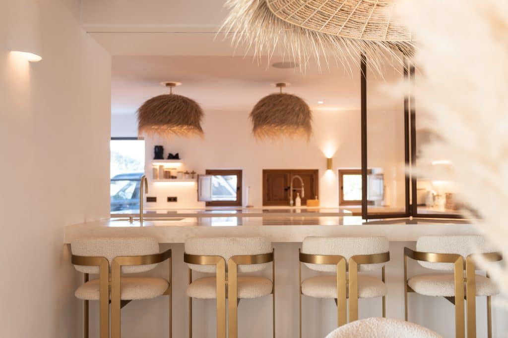 Modern kitchen with a row of four gold and white bar stools at a counter. The background features pendant lights with woven straw shades and warm, neutral-toned décor. | Ibiza