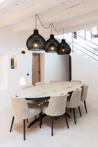 A modern dining area with an oval wooden table surrounded by eight beige upholstered chairs, under three black woven pendant lights. The room has white walls, a wooden ceiling, and a visible staircase. | Ibiza
