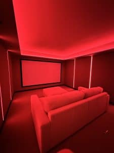 A modern home theatre room with a large screen, a plush sofa, and ambient red lighting along the ceiling and walls, creating a cosy atmosphere. | Ibiza