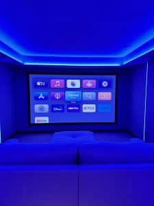 A home theatre room with a large screen displaying various streaming app icons, blue LED lighting, and a comfortable sofa facing the screen. | Ibiza
