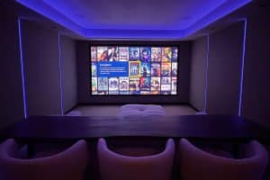 A modern home theatre room with cosy chairs and a large screen displaying a film selection menu, illuminated by blue LED lights along the ceiling and walls. | Ibiza