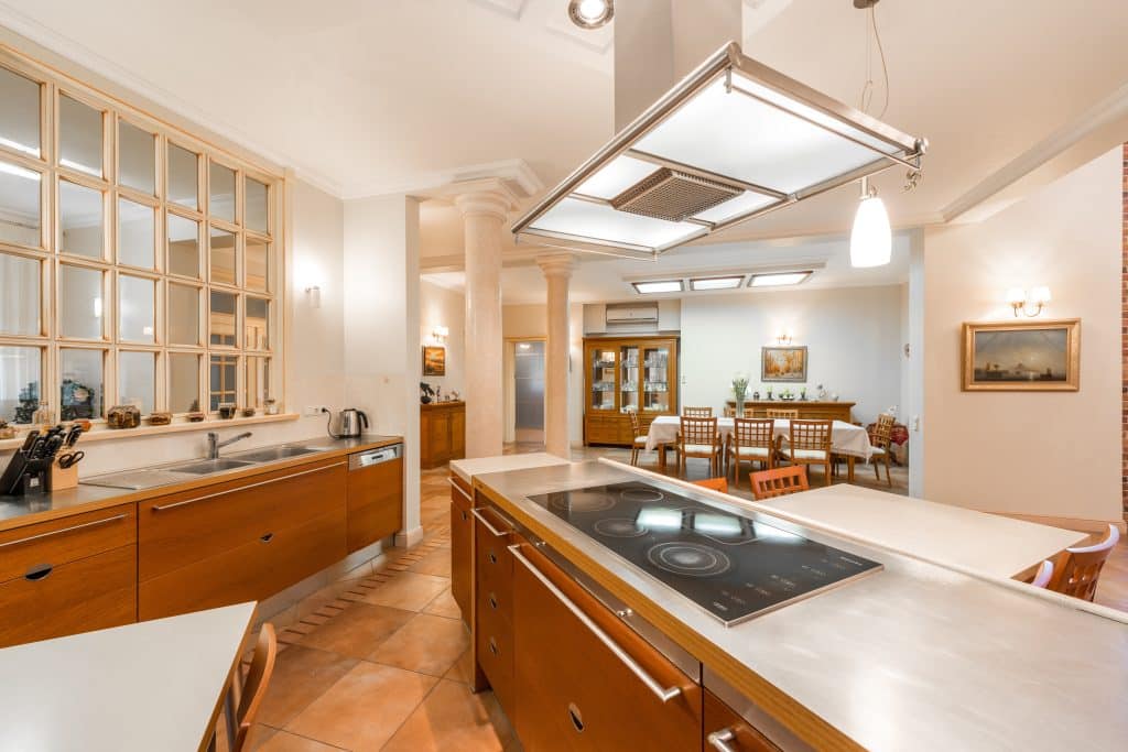 Modern kitchen with wooden cupboards, stainless steel worktops, an electric hob on an island, and an open dining area with a wooden table and chairs in the background. Warm lighting brightens the spacious room. | Ibiza