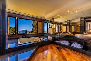 Luxury bathroom with a sunken bath by large windows overlooking greenery and blue sky, double basins, mirrors, wooden floor, and neatly folded towels on open shelves below the counter. | Ibiza
