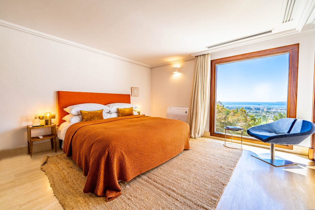 A bright hotel room with a large orange bed, two bedside tables with lamps, a blue modern chair, a large window with a scenic view of trees and the sea, and natural light filling the space. | Ibiza