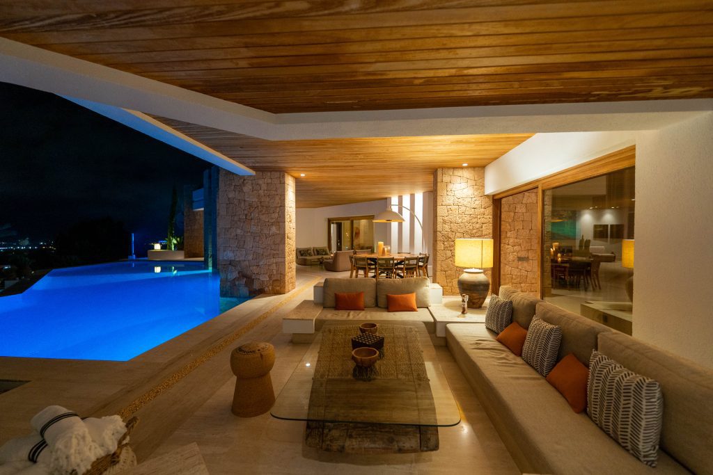 Open-air luxury patio at night with a modern pool glowing blue, cosy cushioned seating, a glass coffee table, and warm ambient lighting. Stone and wood elements create an inviting, elegant atmosphere. | Ibiza