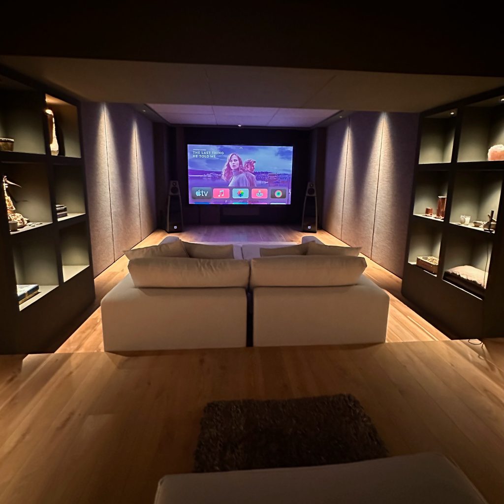 A modern home theatre room with a large screen displaying a streaming interface, two white sofas, wooden floors, and built-in shelves with decor along the walls. The room has dim lighting and a cosy atmosphere. | Ibiza