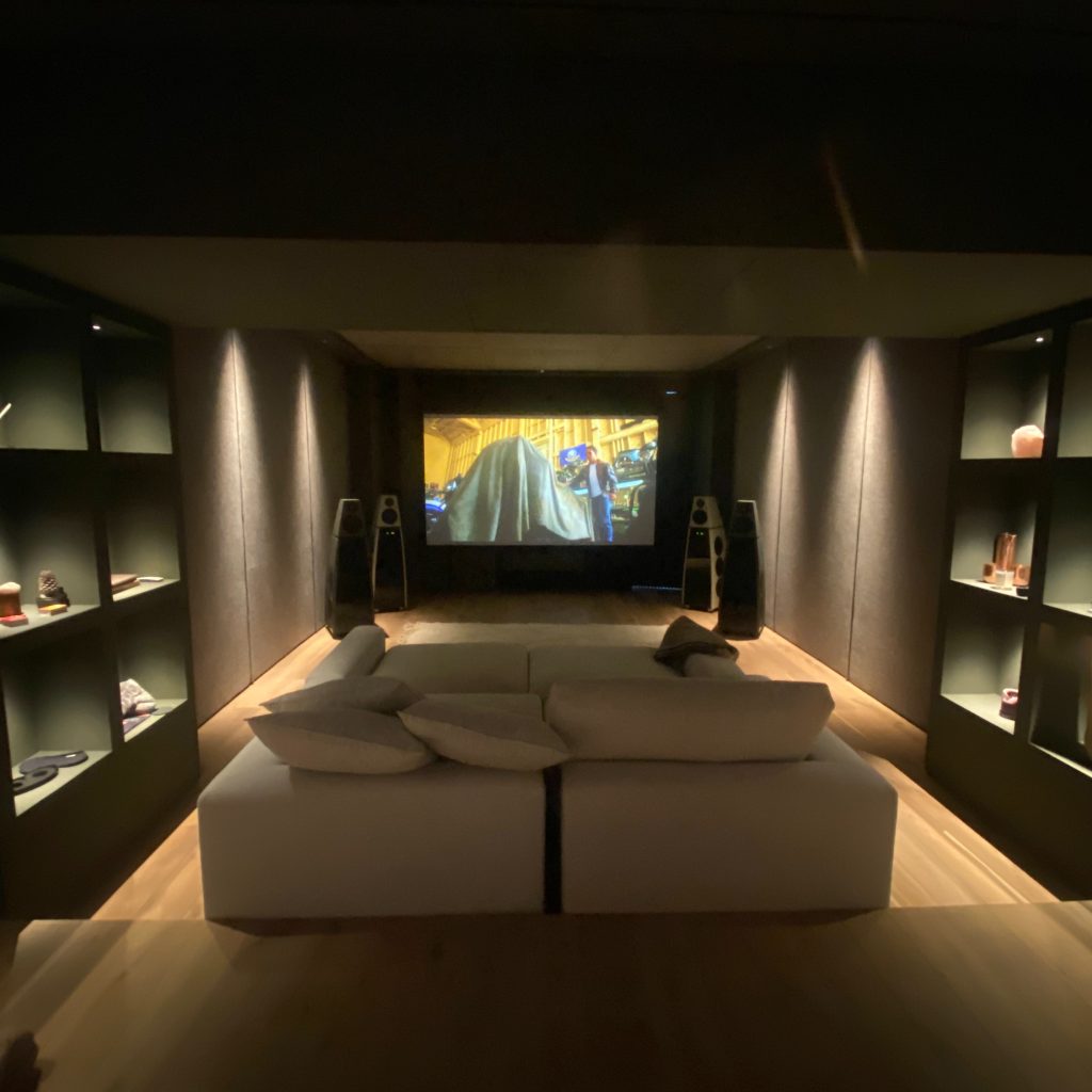 A cosy home theatre room with a large screen displaying a film, two speakers on either side, shelves with decorations along the walls, and a beige sofa with cushions facing the screen. | Ibiza