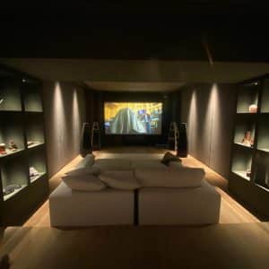 A cosy home theatre room with a large screen displaying a film, two speakers on either side, shelves with decorations along the walls, and a beige sofa with cushions facing the screen. | Ibiza