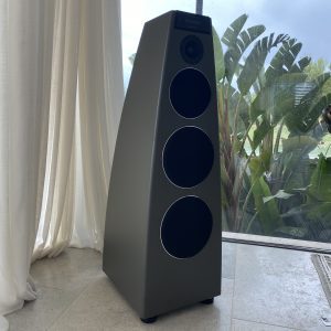 A tall, modern black speaker with three large circular drivers stands on a tiled floor near a sheer curtain and large window with tropical plants visible outside. | Ibiza