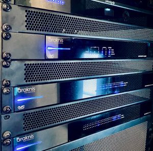 A close-up of several stacked black server units with glowing blue indicator lights in a network rack, displaying the brand name Araknis on the front panels. | Ibiza