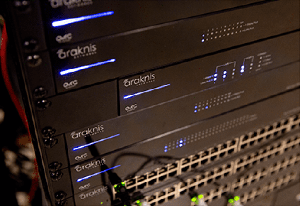 Close-up of several Araknis network servers and switches stacked in a server rack, with blue indicator lights illuminated and multiple Ethernet ports visible. | Ibiza