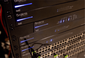 Close-up of several Araknis network servers and switches stacked in a server rack, with blue indicator lights illuminated and multiple Ethernet ports visible. | Ibiza