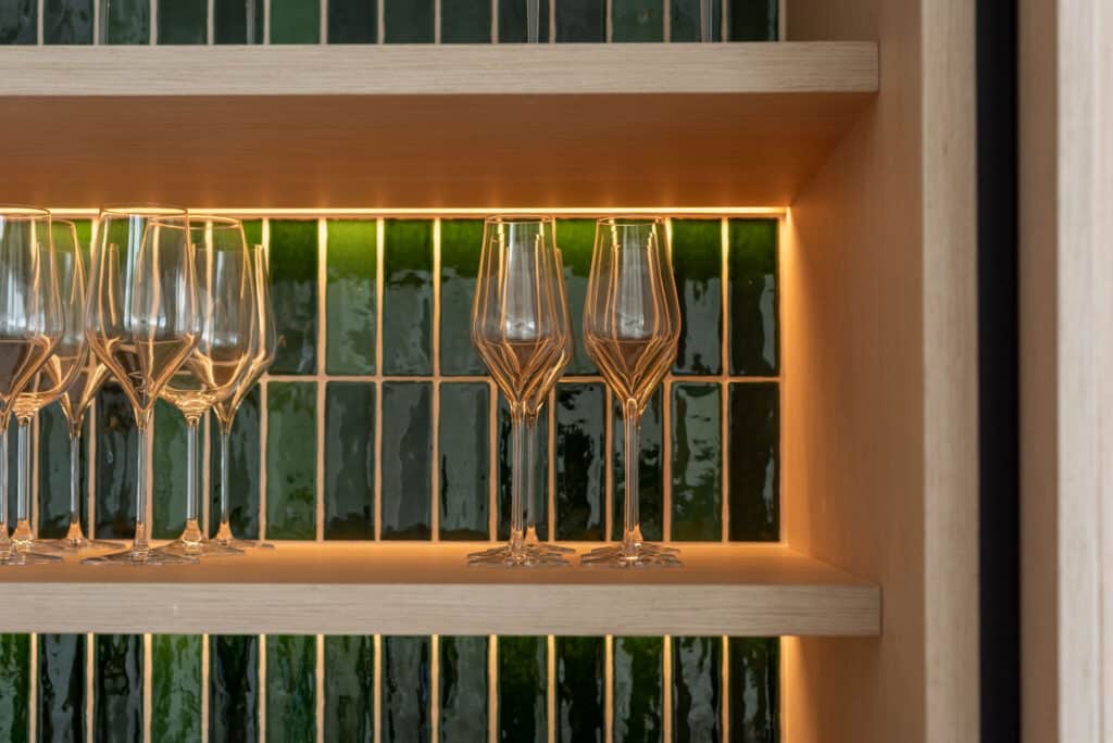 A wooden shelf with wine and cocktail glasses neatly arranged, illuminated by warm lighting, adds a cosy touch to this Talamanca flat. The background features glossy green rectangular tiles arranged vertically. | Ibiza