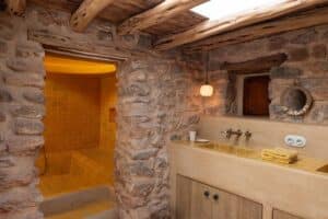 Rustic bathroom with stone walls, exposed wooden beams, a tiled shower area, a beige vanity with yellow tiles, a round mirror, a small window, and folded yellow towels on the worktop. Warm, cosy lighting throughout. | Ibiza