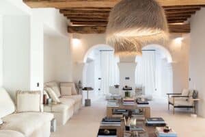 Bright living room with white walls, exposed wooden ceiling beams, two large archways, neutral-toned sofas, stacked coffee tables with books, and two large pendant lights with a natural, woven texture. | Ibiza
