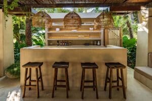 A cosy outdoor bar with four wooden stools, hanging woven pendant lights, a simple counter, and shelves holding bottles and glassware, surrounded by lush green tropical plants. | Ibiza