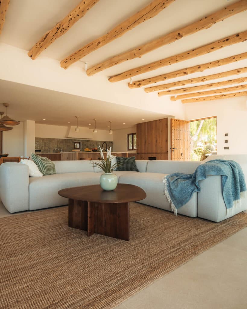A spacious, modern living room with a light grey sectional sofa, a wooden coffee table, a woven rug, and exposed wooden ceiling beams. A blue throw blanket and a vase with flowers decorate the space. The kitchen is visible in the background. | Ibiza