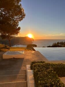 Sunset over the ocean viewed from the stone terrace of Casa Enchanted, with an infinity pool, covered lounge bed, manicured shrubs, and tall trees. Warm sunlight reflects on the water and cliffs in the background. | Ibiza