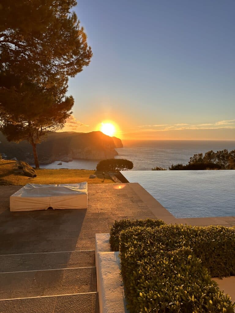 Sunset over the ocean viewed from the stone terrace of Casa Enchanted, with an infinity pool, covered lounge bed, manicured shrubs, and tall trees. Warm sunlight reflects on the water and cliffs in the background. | Ibiza