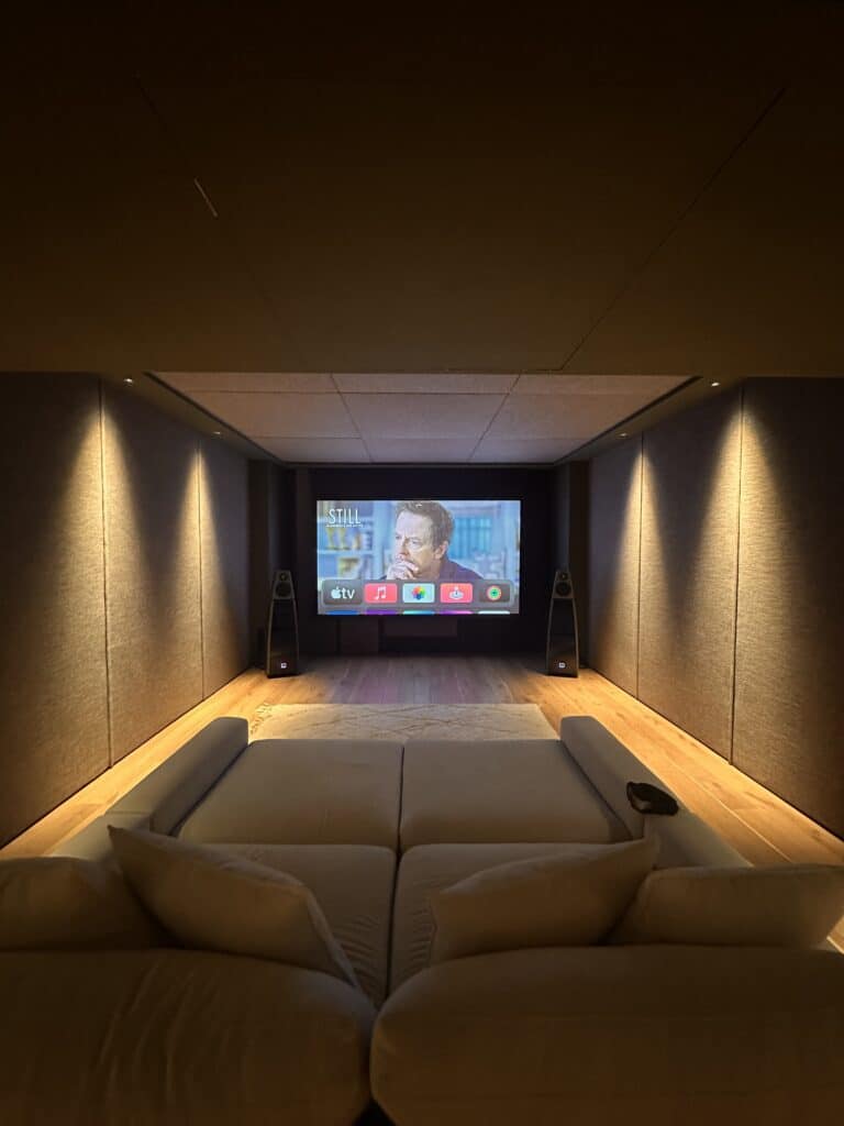 A cosy home theatre room with a large screen displaying a streaming menu, a man’s face on the screen, surround speakers, soft lighting, and a comfortable light-coloured sofa in the foreground. | Ibiza