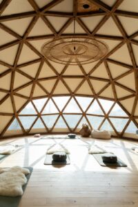 A spacious, light-filled geodesic dome with wooden framework features yoga mats, cushions, and sheepskin rugs arranged on a wooden floor for meditation or yoga practice. Large windows offer views of a natural landscape outside. | Ibiza