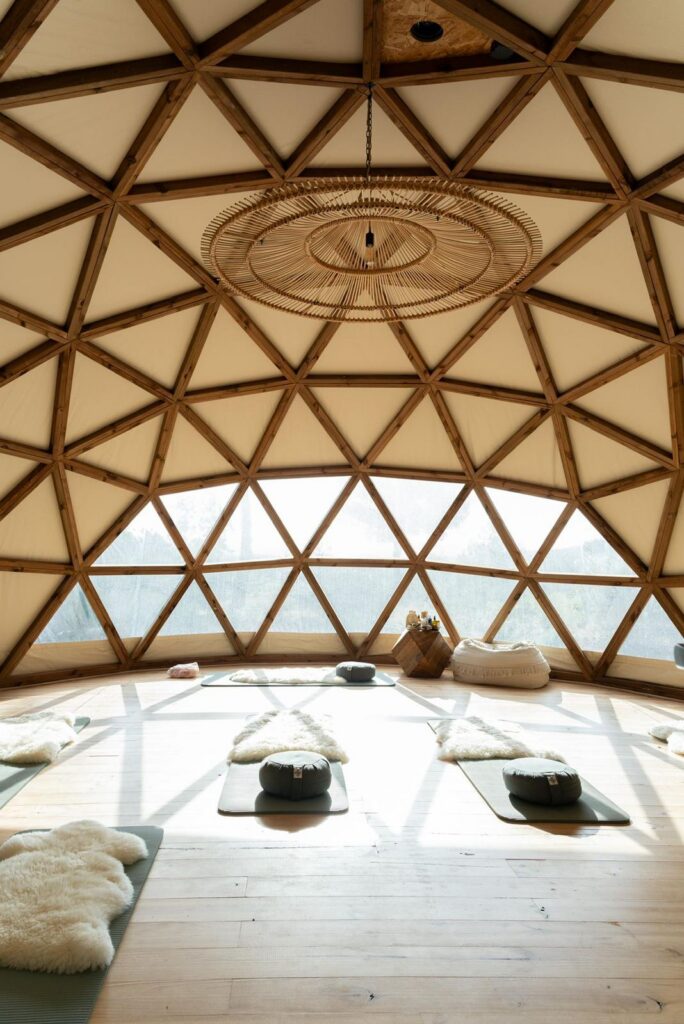 A spacious, light-filled geodesic dome with wooden framework features yoga mats, cushions, and sheepskin rugs arranged on a wooden floor for meditation or yoga practice. Large windows offer views of a natural landscape outside. | Ibiza