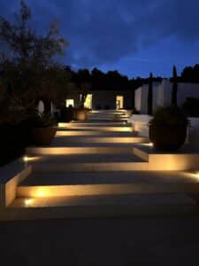 Wide stone steps lit by warm, recessed lights lead up to a modern house at night, with large potted plants on either side and trees silhouetted against a dark, cloudy sky. | Ibiza