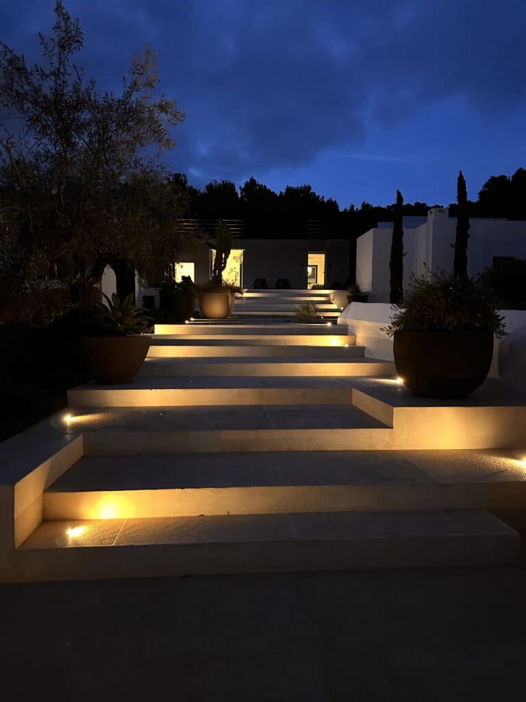 Wide stone steps lit by warm, recessed lights lead up to a modern house at night, with large potted plants on either side and trees silhouetted against a dark, cloudy sky. | Ibiza