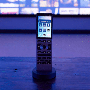 A modern remote control with a touchscreen display stands upright on a dock, with a blurred TV screen showing film thumbnails in the background. The scene is lit with a blue glow. | Ibiza